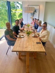 group_of_people_sitting_around_long_wooden_table_with_food_and_flowers_in_bright_room_with_large_window_view